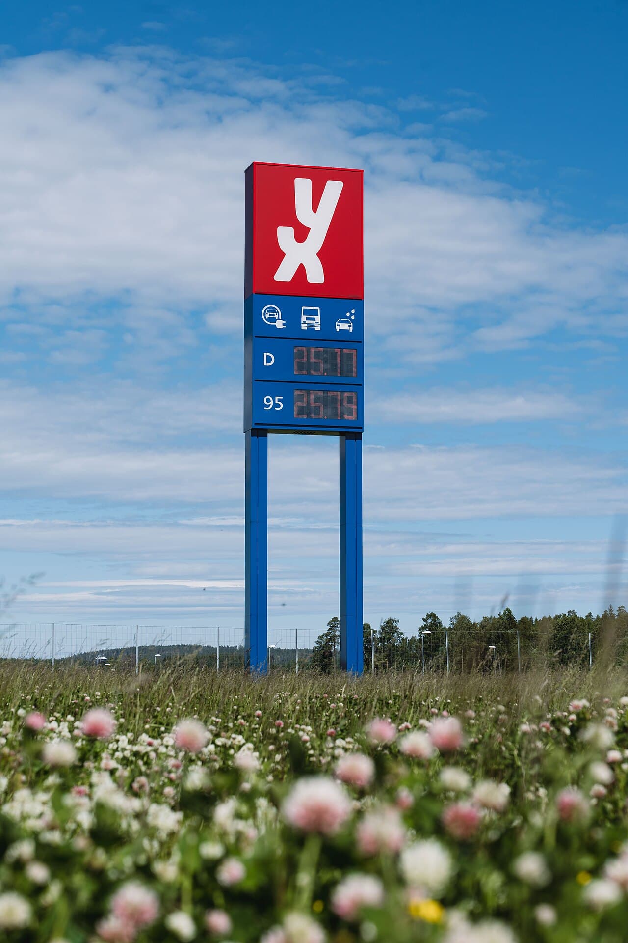 Høyt mastskilt med rød YX-logo og prisdisplay mot blå himmel, med blomstereng i forgrunnen.