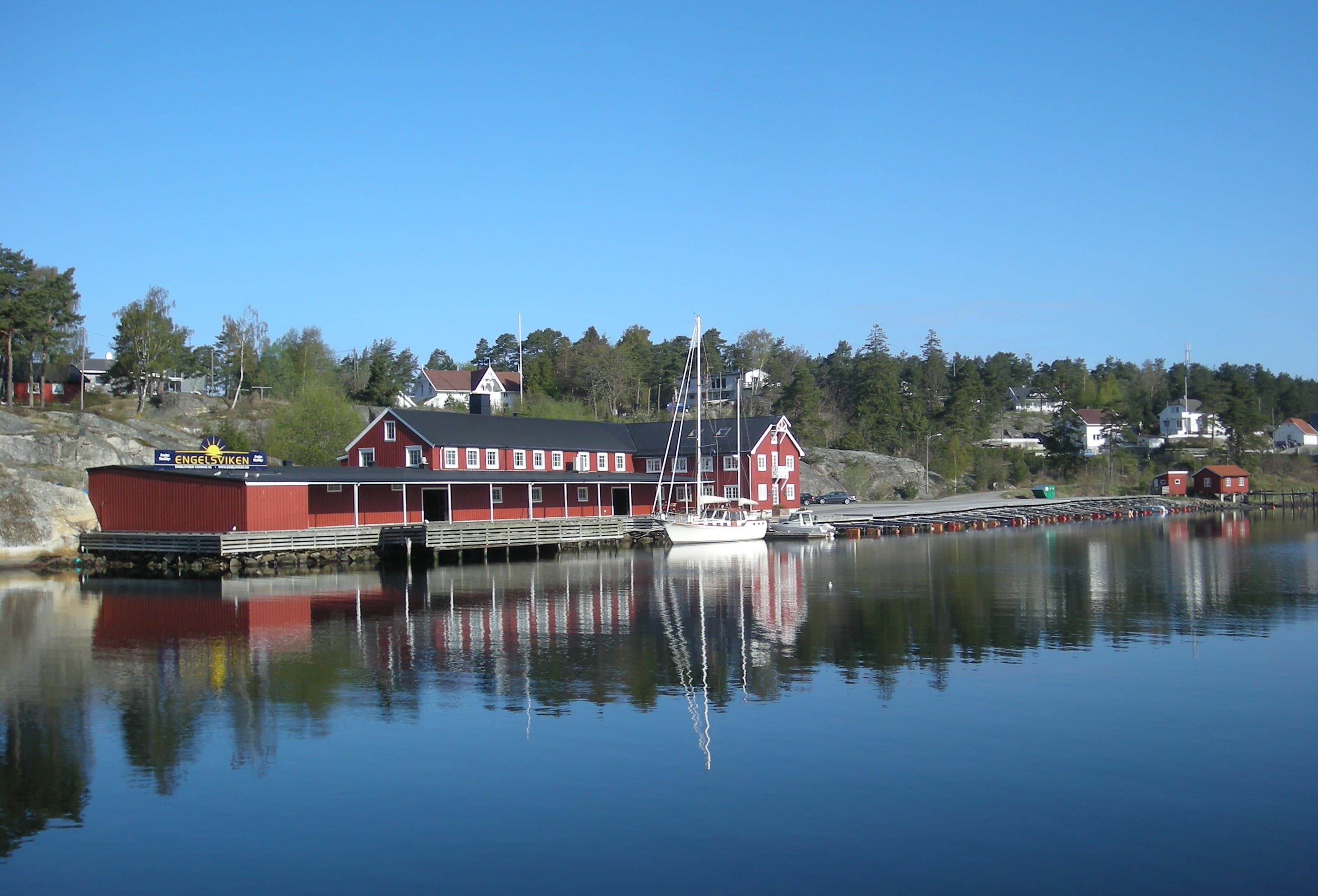 Engelsviken havn med lange røde sjøboder, en hvit seilbåt ved en brygge og en steinmolo speilet i stille vann.